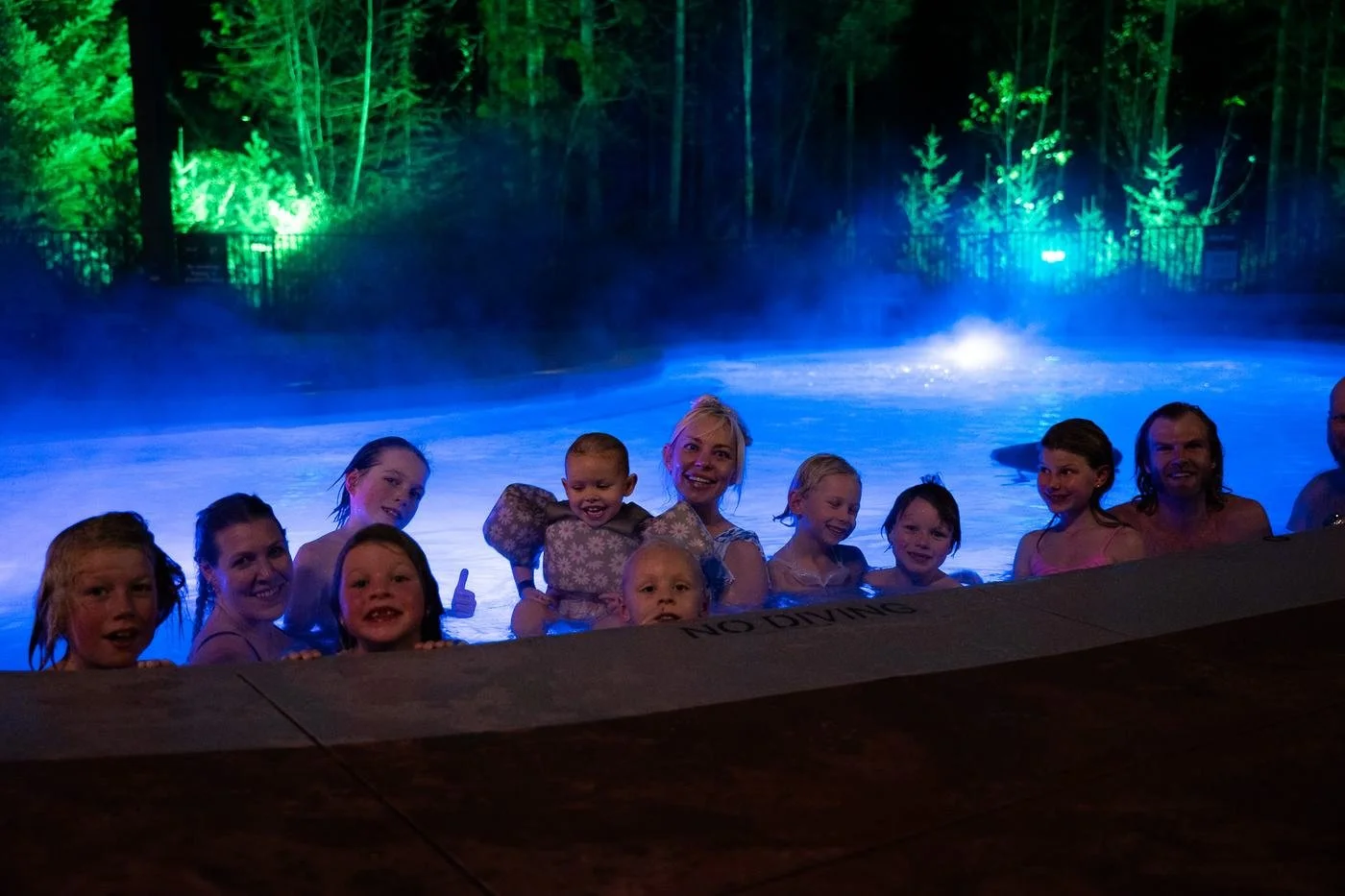 A large family group smiling together in a blue-lit hot pool at night