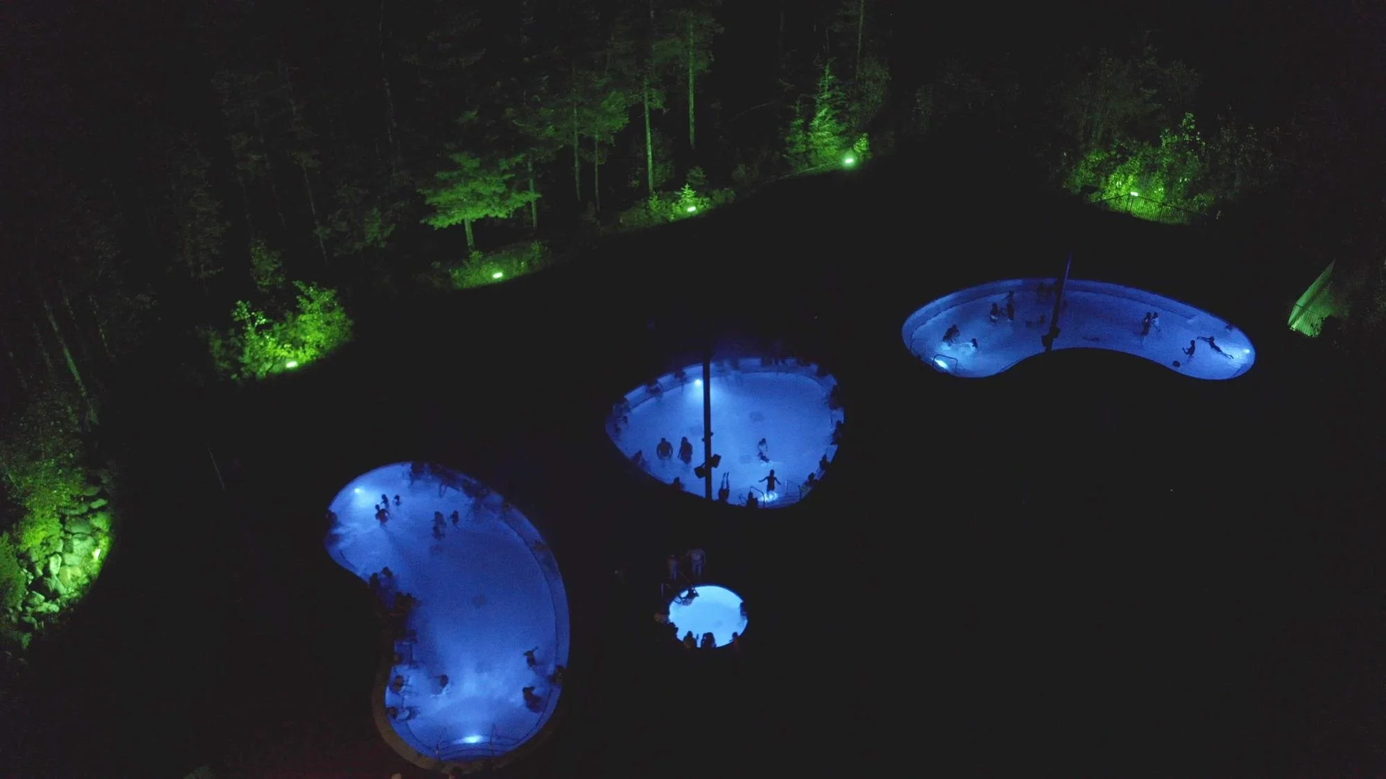 Drone view at night of Crazy Creek's three large pools and round cold plunge, lit blue, surrounded by forest lit green
