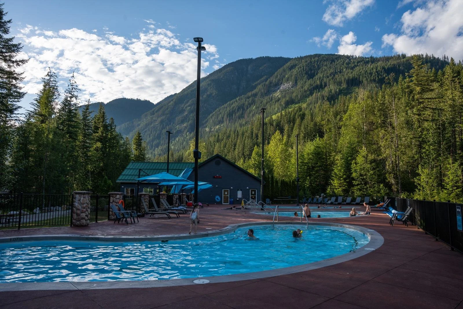 Guests soaking in a sunny outdoor pool at Crazy Creek, with forested mountains and blue sky behind
