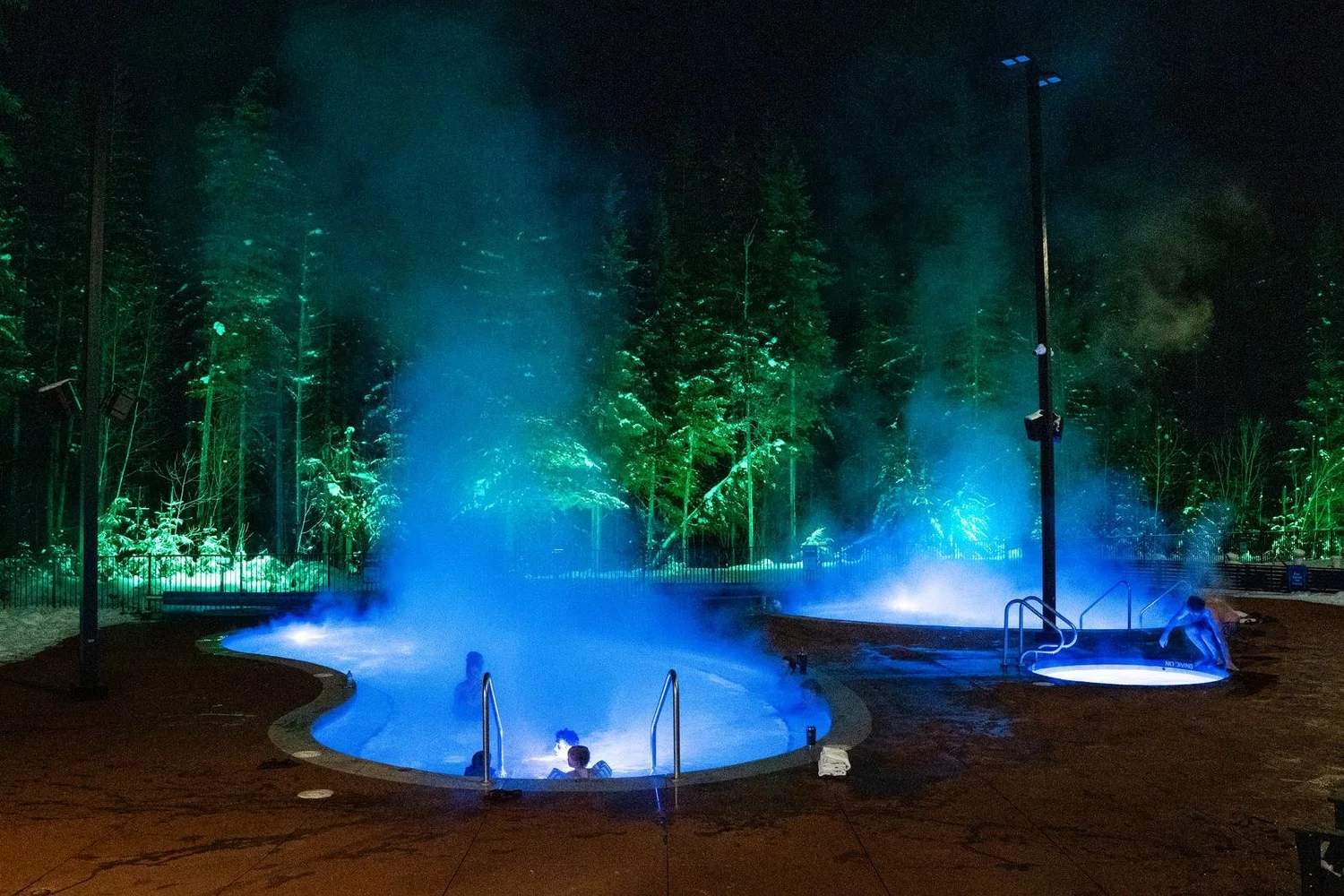 Steam rising off the blue-lit Crazy Creek hot pools on a winter night, with snowy trees lit green behind