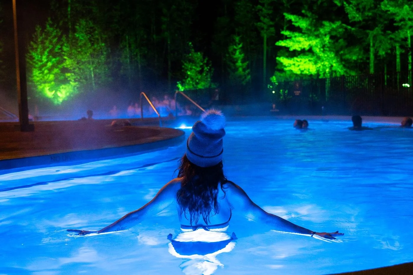 A woman in a knit toque floating with her arms spread in a blue-lit pool at night, forest lit green behind