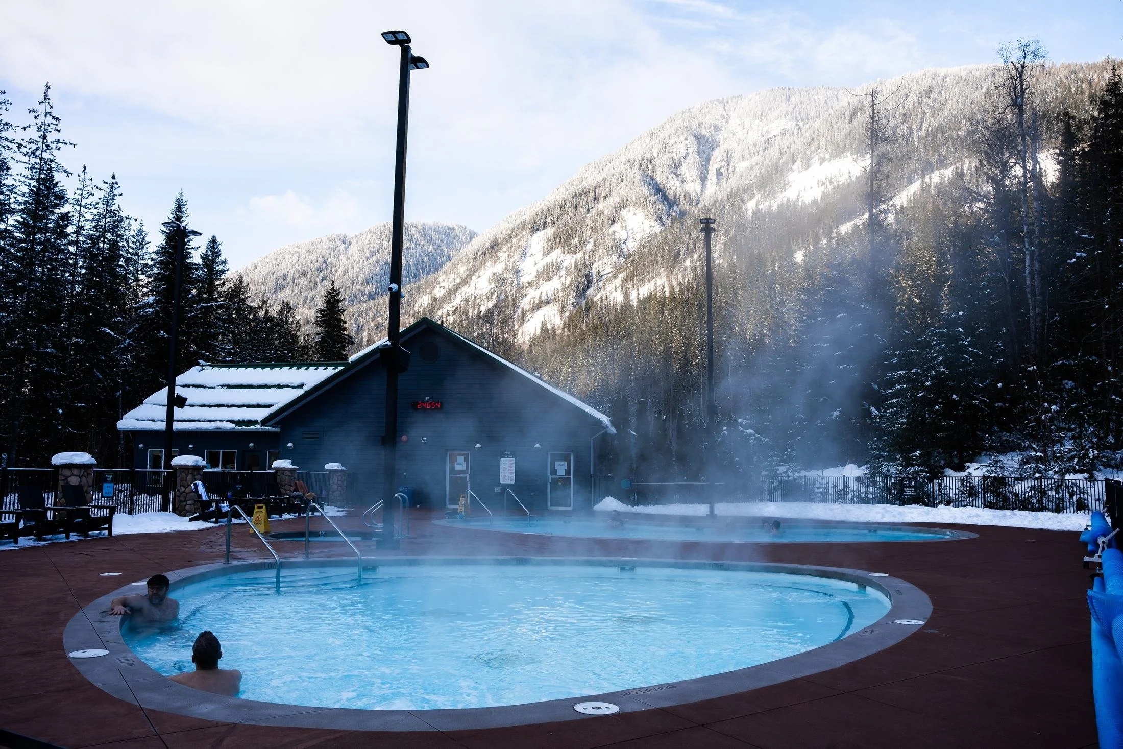 Steam rising off an outdoor hot pool at Crazy Creek on a winter morning, with the pool building and snowy mountains behind