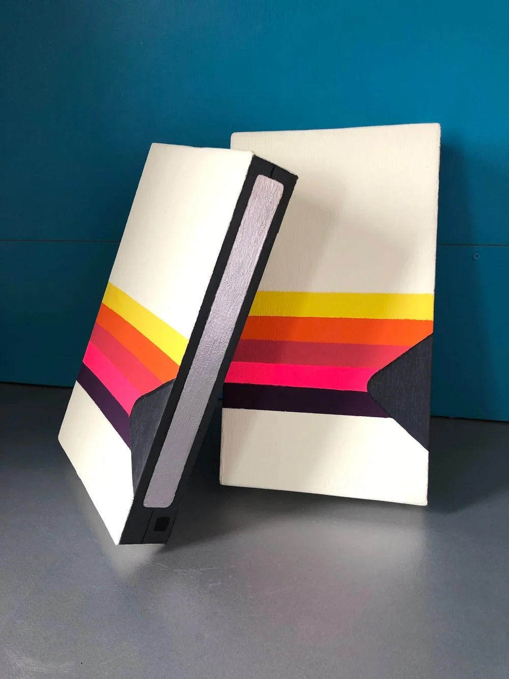 Blank Tape, two painted pieces in the form of VHS tapes with rainbow stripes, leaning against a teal wall