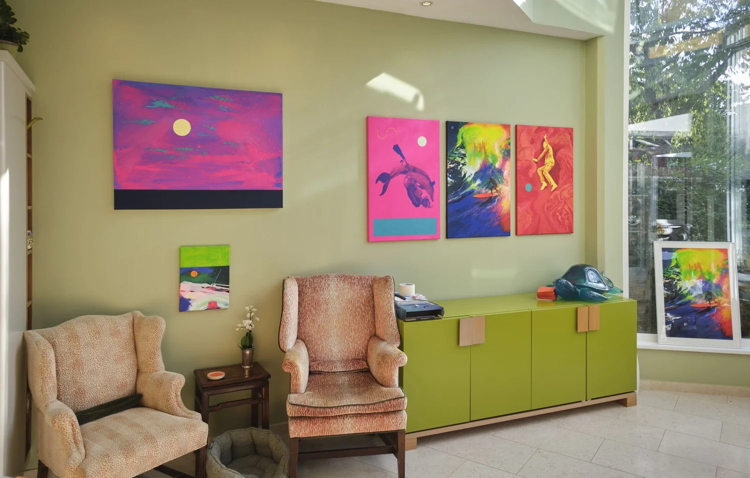 Paintings hung in a sunlit room with two armchairs