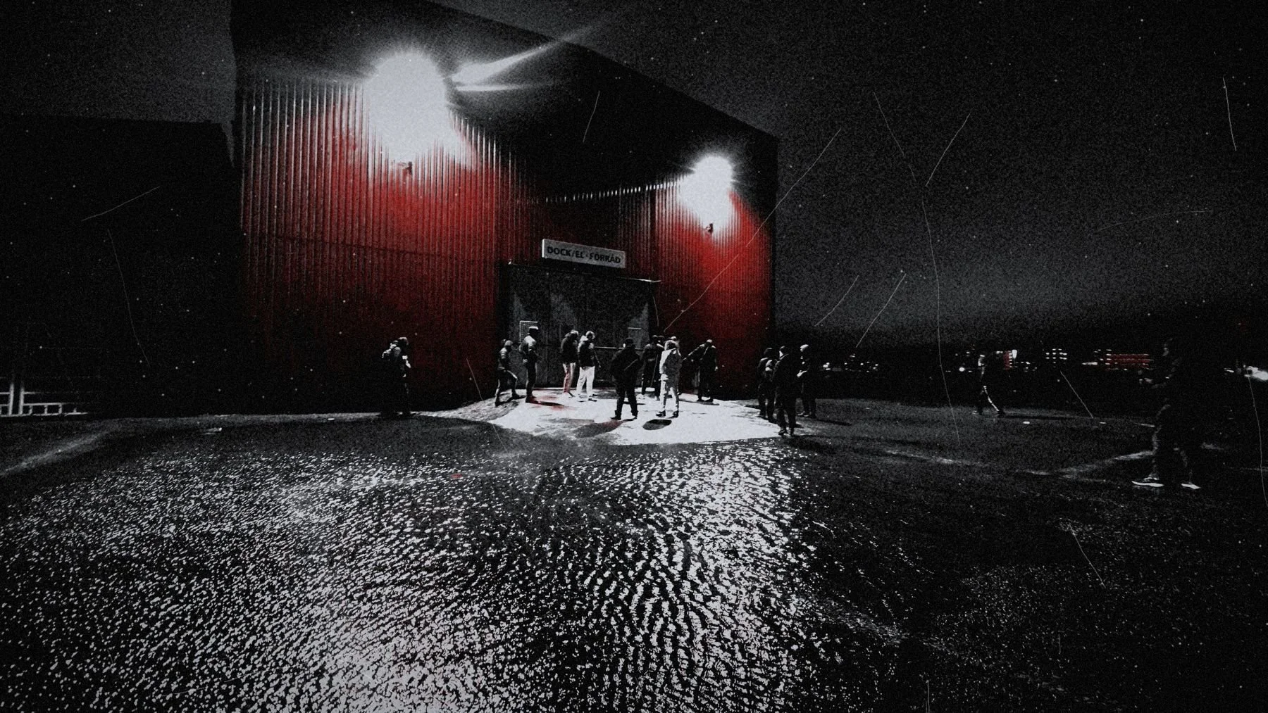 A red warehouse lit by floodlights at night, people at its door on wet ground