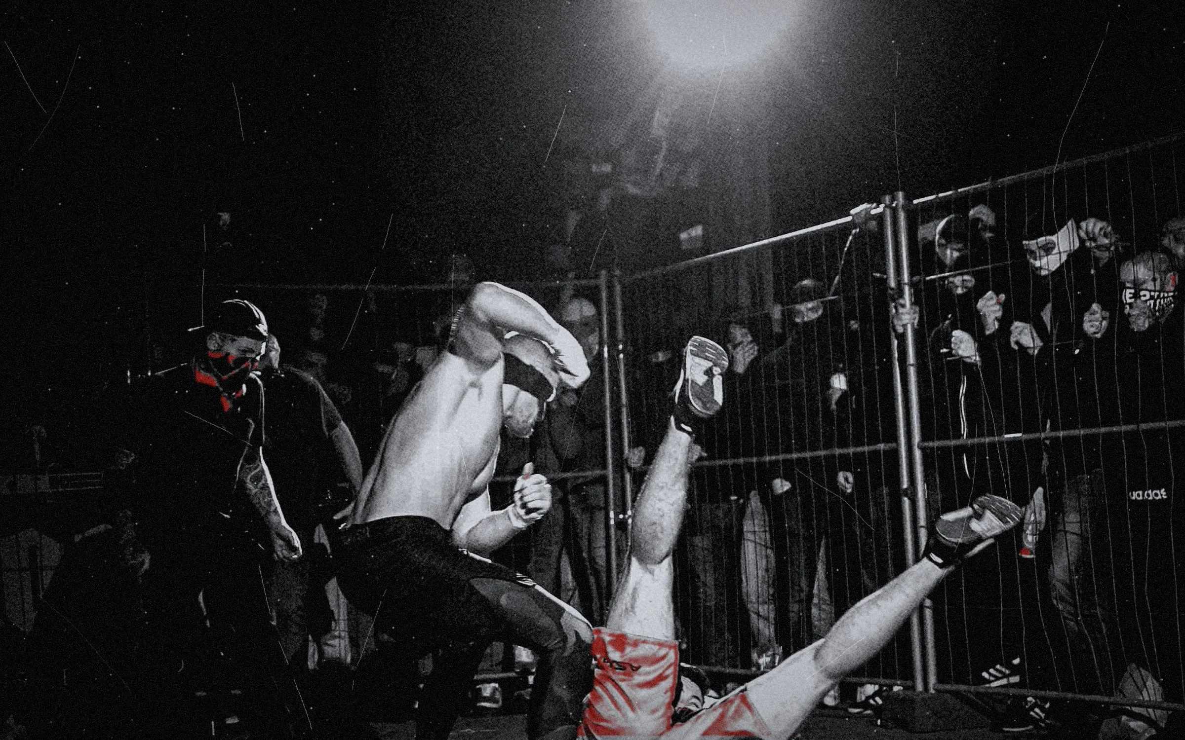 A shirtless fighter kicking his opponent, who flips upside down under a floodlight, masked crowd at the fence