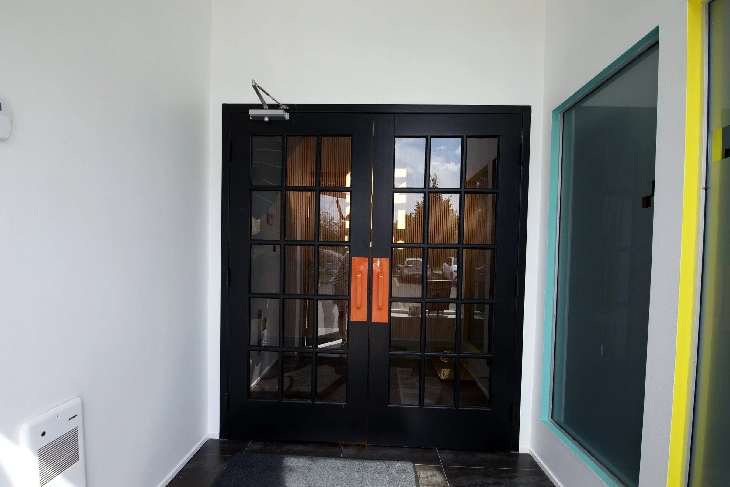 After, August 2024: the same entrance doors, now black with orange handles. This photo was scaled and shifted to line up with the before photo; the only other change is a blurred reflection in the door glass.