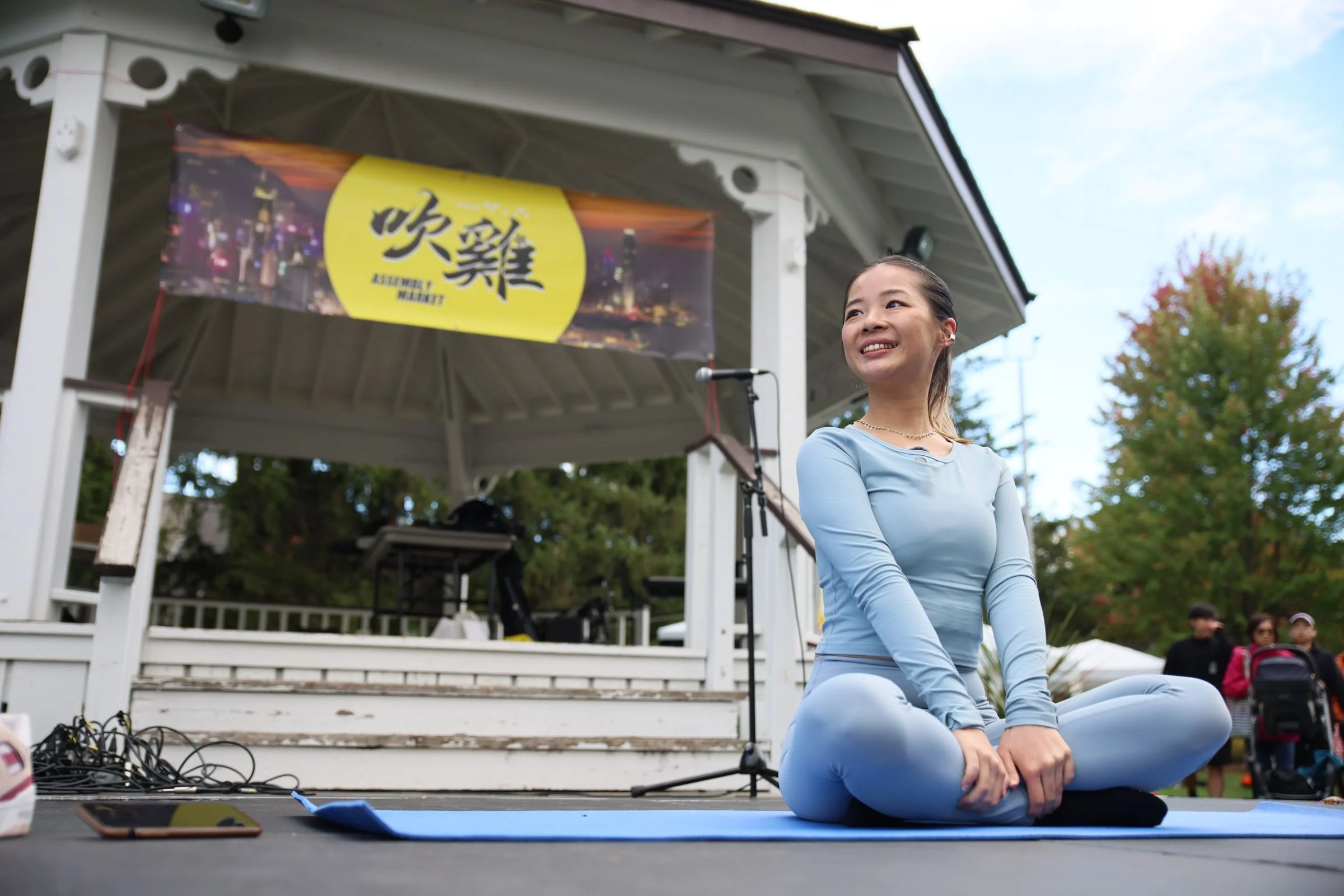 A yoga teacher in light blue sits on a mat in front of the bandstand, smiling, under the yellow 吹雞 stage banner.