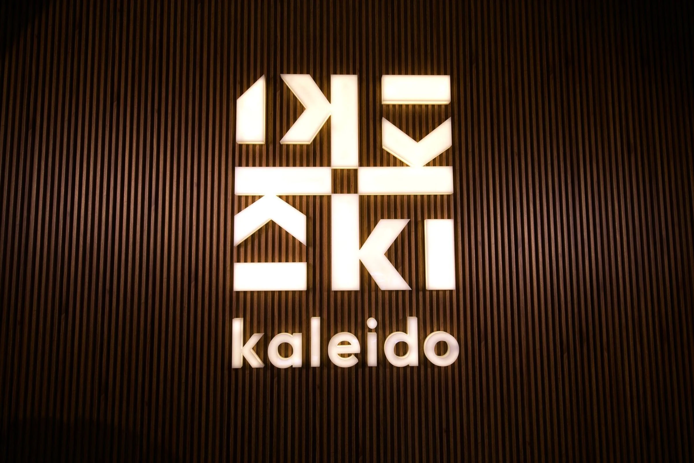 The lit Kaleido logo on a dark wood slat wall inside the finished venue, August 2024.