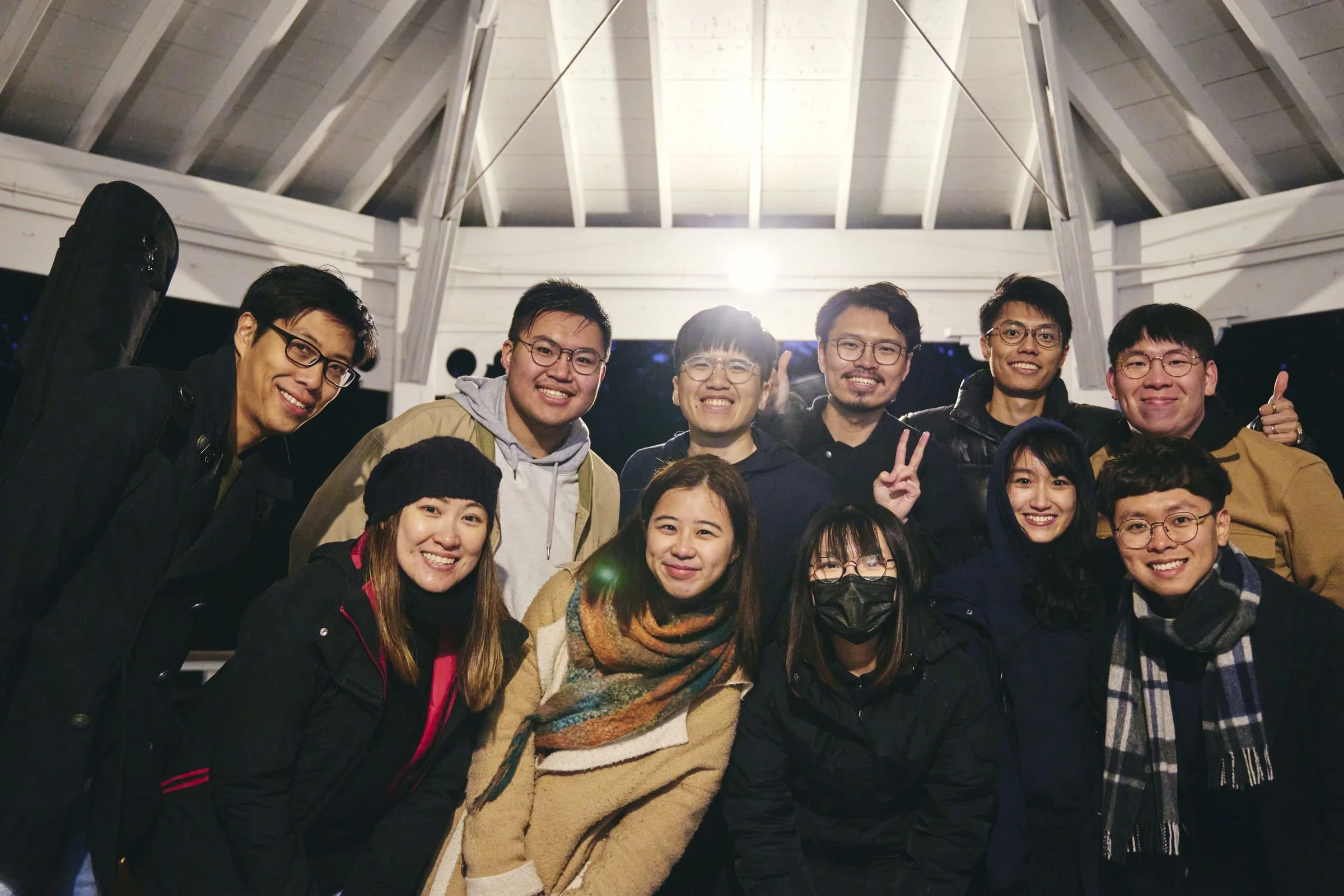 A group of about fifteen people in coats and scarves smile together under the bandstand roof at night.