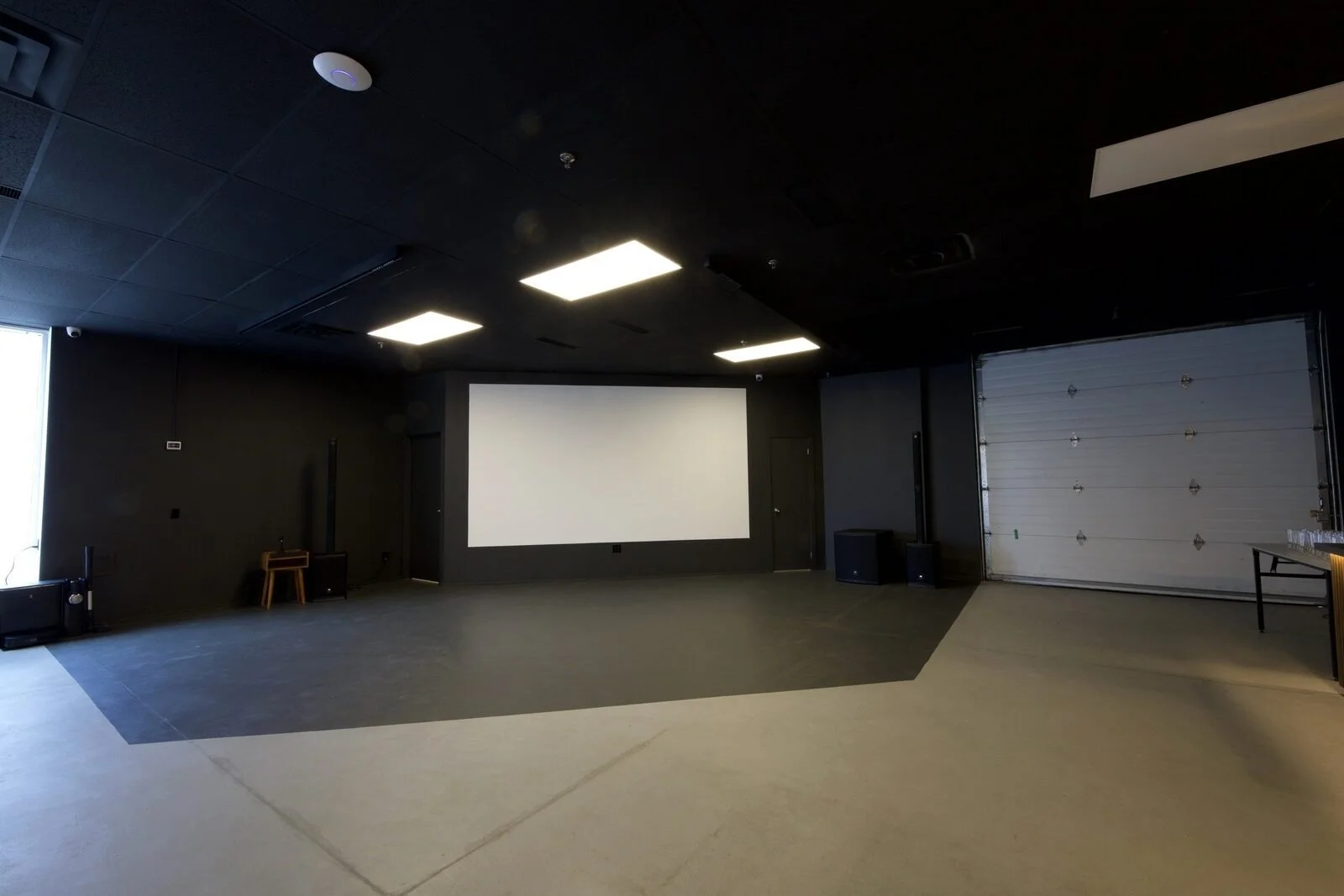 After, August 2024: the hall seen wide, with a black ceiling, a projection screen and the same garage door on the right.