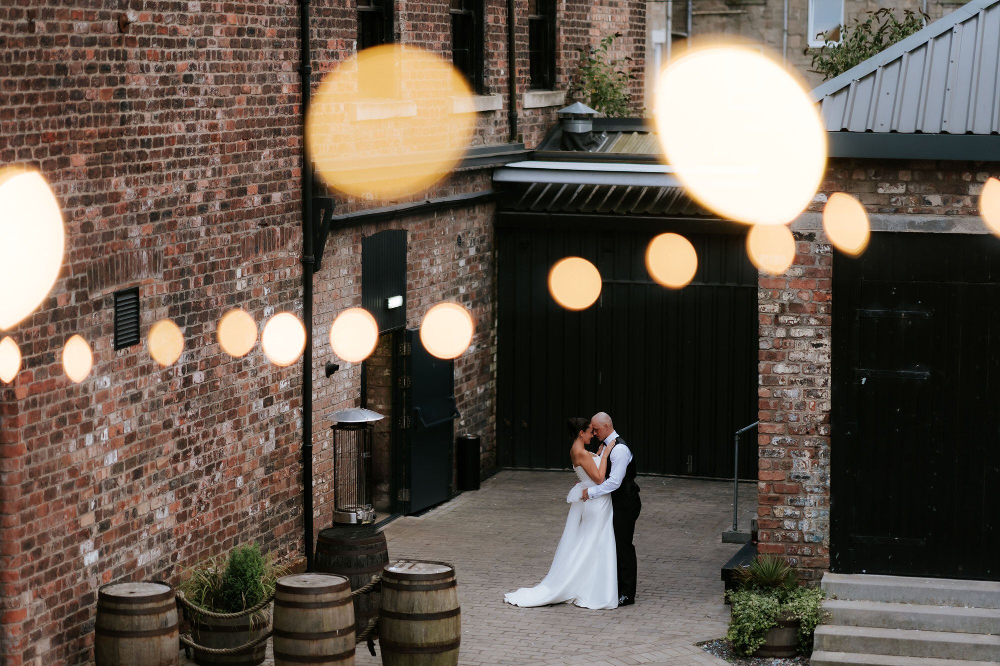 Caitlin and Craig embracing beneath glowing festoon lights in the brick courtyard at The Engine Works.