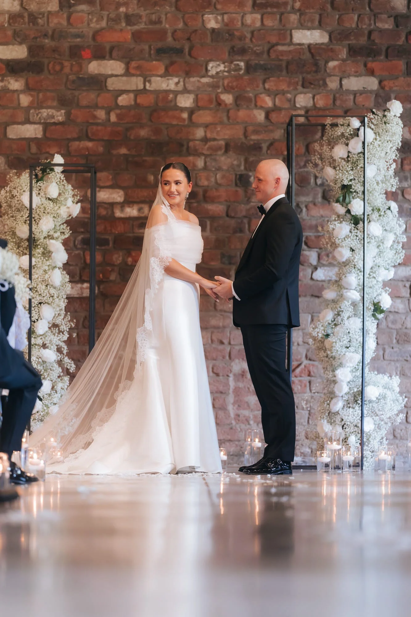 Caitlin and Craig holding hands during their ceremony between white floral arrangements.