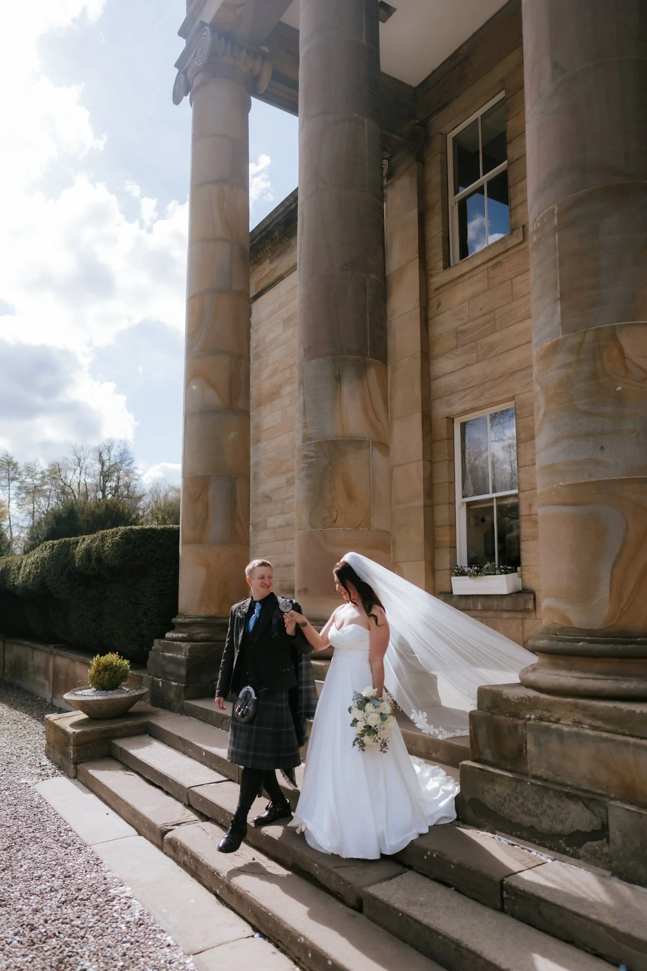 Steph and Calum standing on the sunlit steps at Balbirnie House.