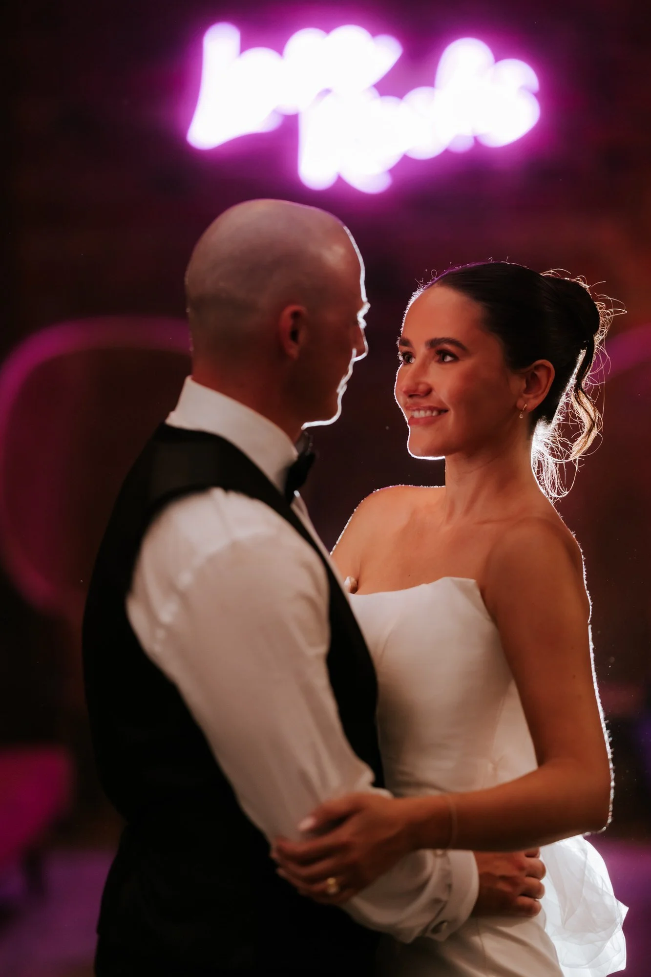 Caitlin smiling at Craig in the glow of the pink neon sign.