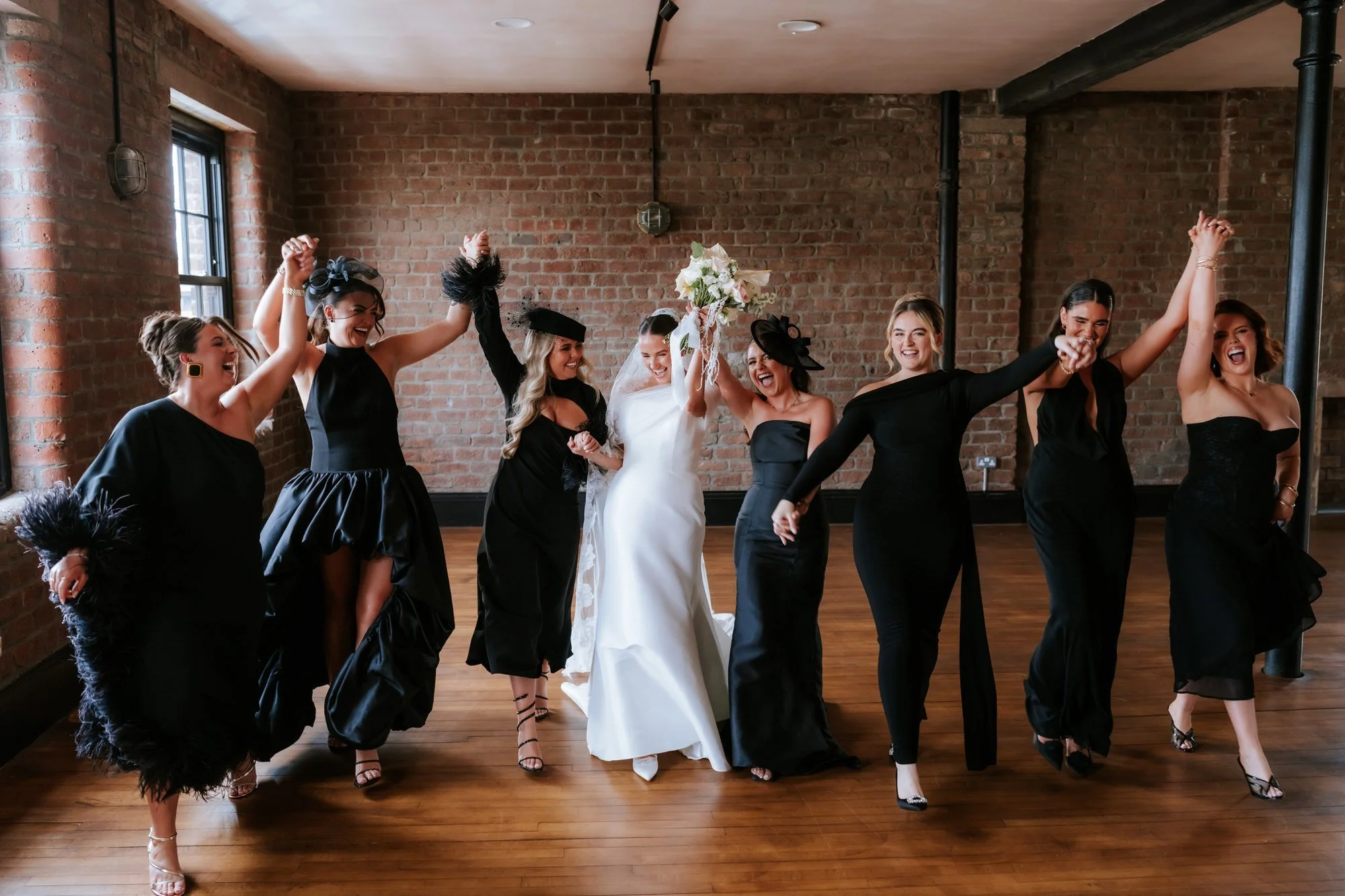Caitlin raising her bouquet with friends in black dresses inside The Engine Works.