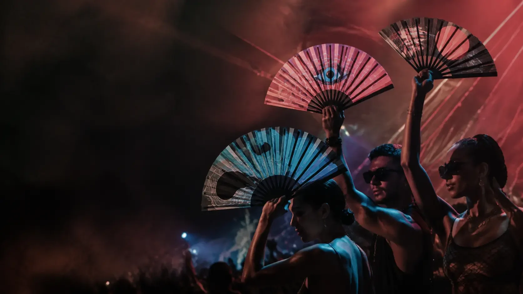 Three festival fans raised beneath coral and cyan lights at an electronic music event