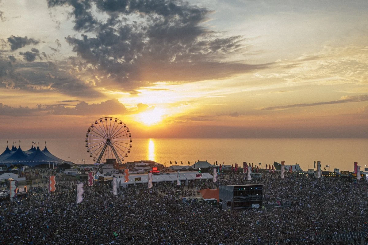 Elevated view over a large festival site at sunset, with crowds, stages and a Ferris wheel