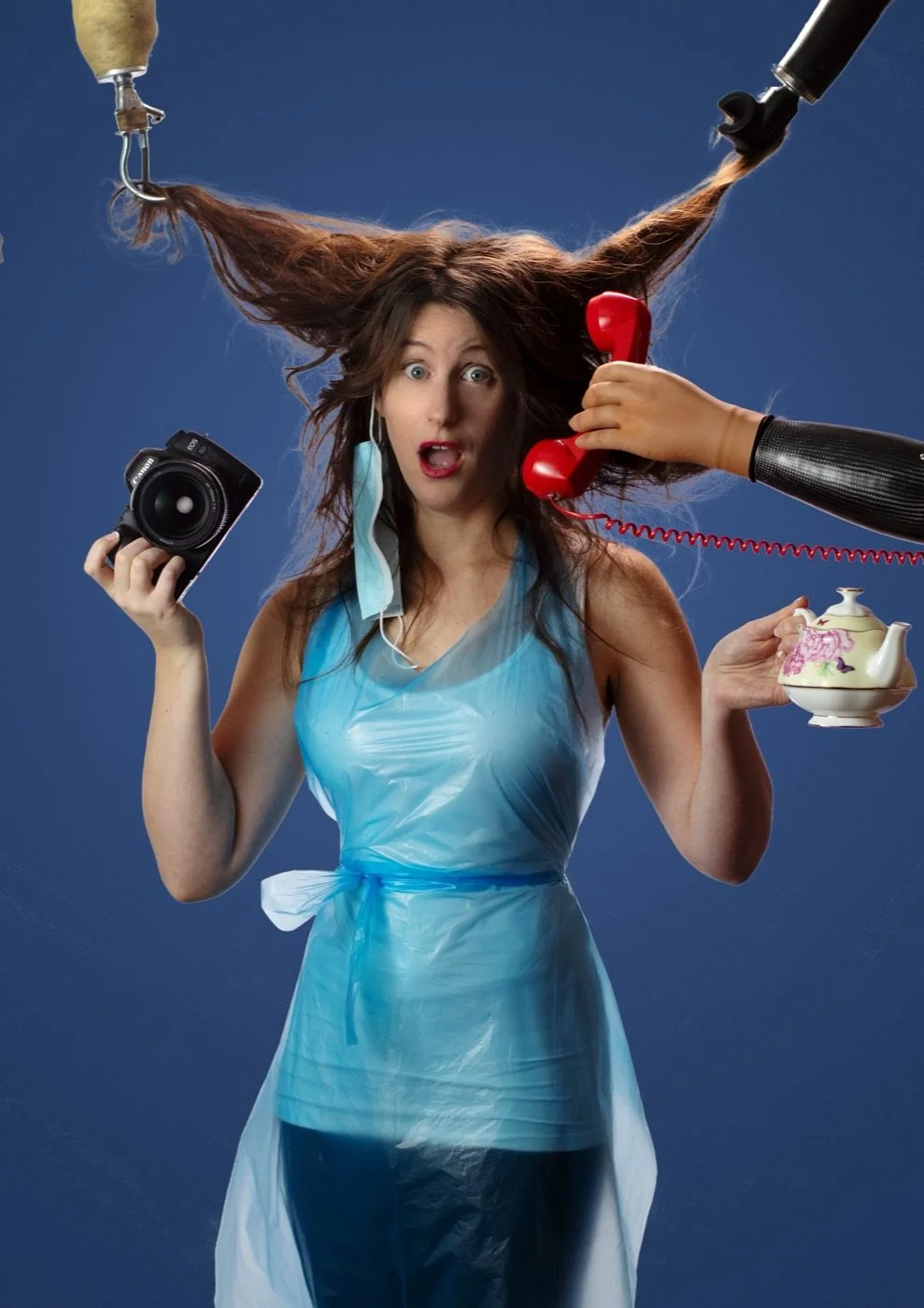 Jennifer in a plastic apron, hair pulled up by hooks, holding a camera and a teapot