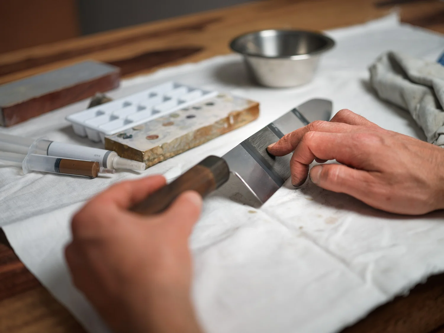 Two hands finishing a blade on a bench stone, finger stones and slurry on the towel beside it
