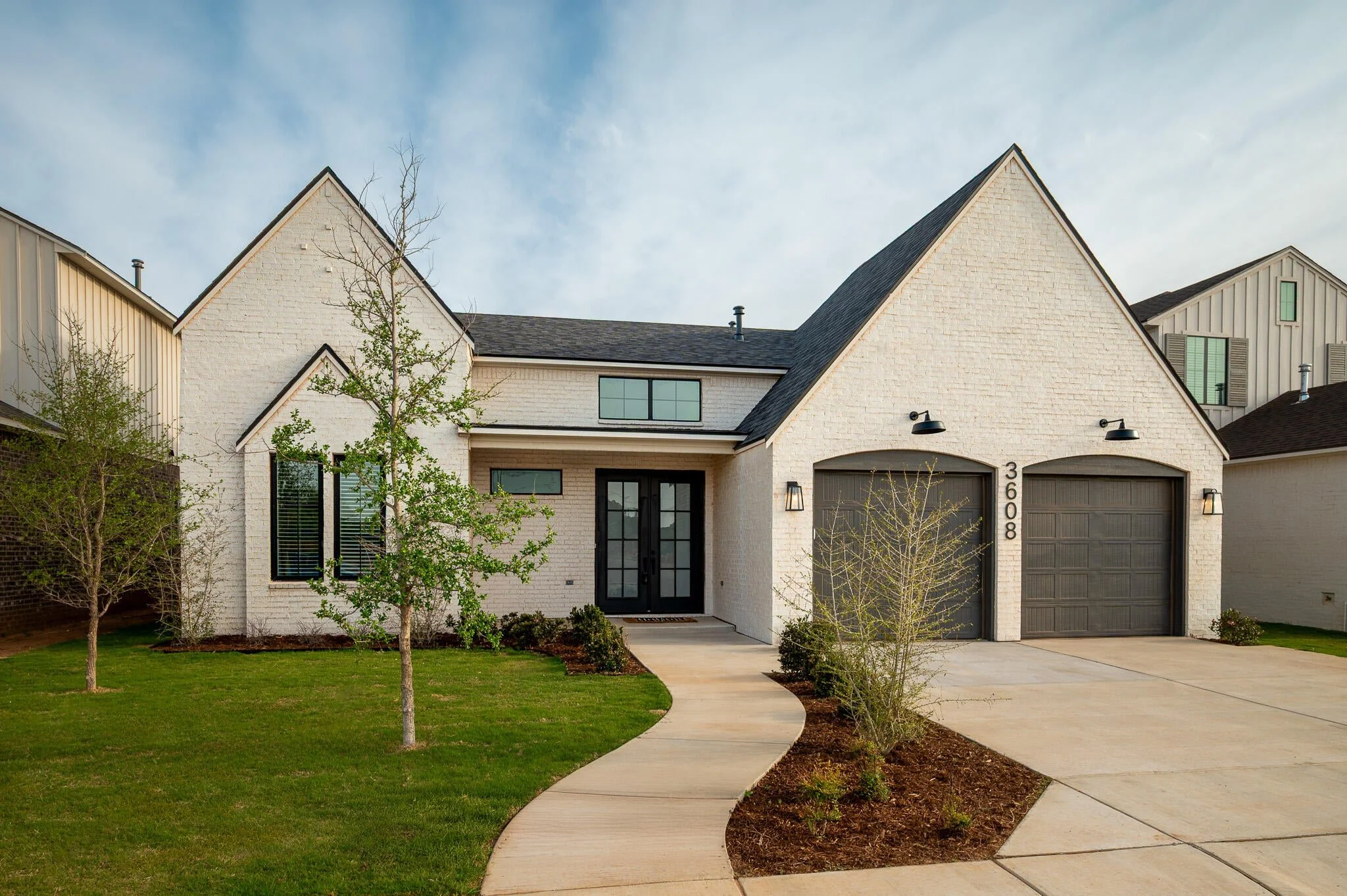 White brick home with black-framed windows, a landscaped front walk, and two garage doors.