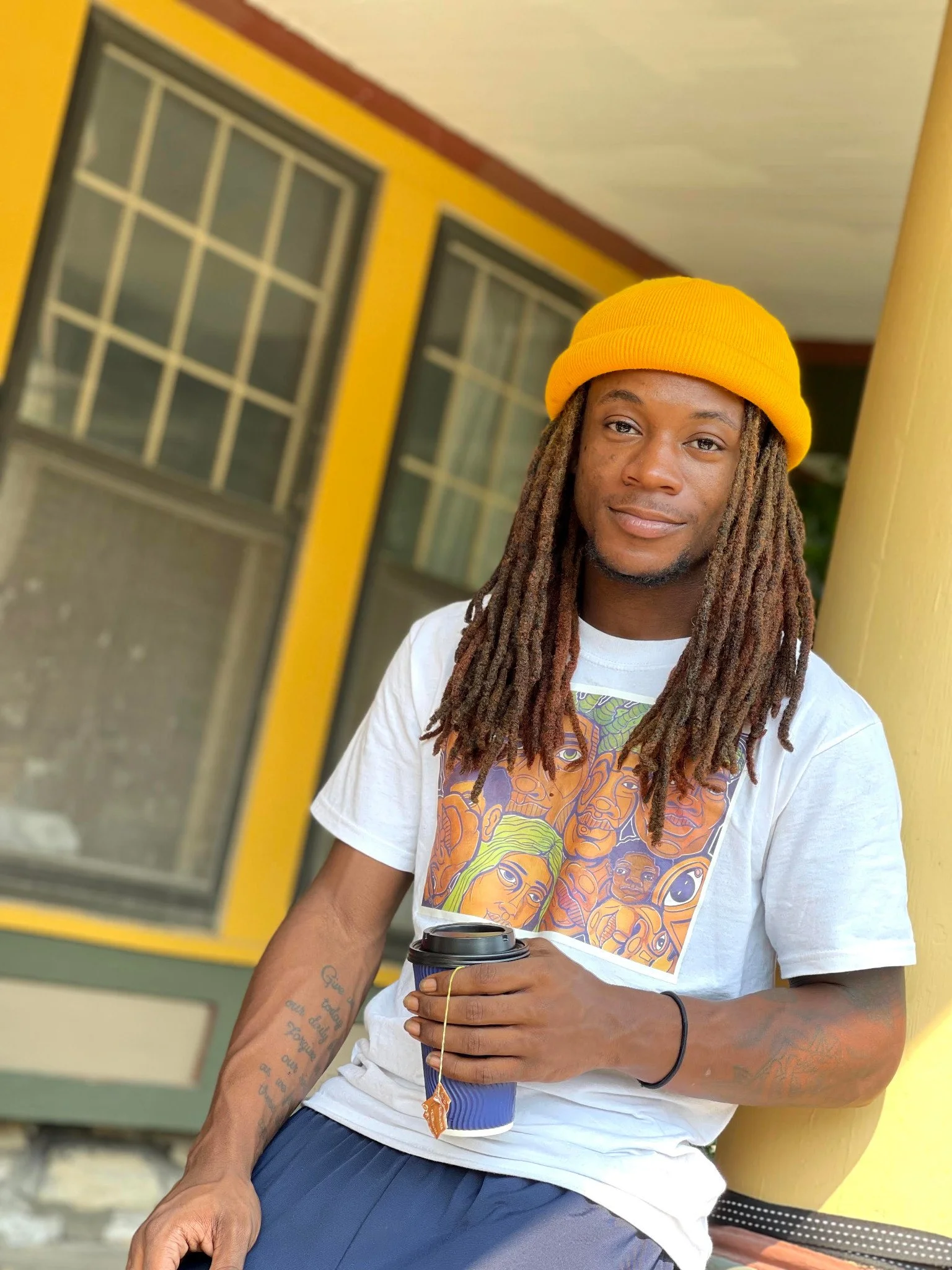 Portrait of a person in a yellow beanie holding a cup on a porch.