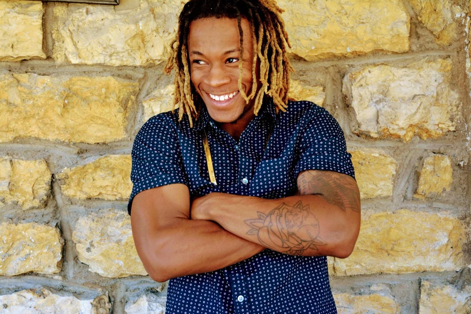 Smiling portrait in a patterned shirt against a stone wall.