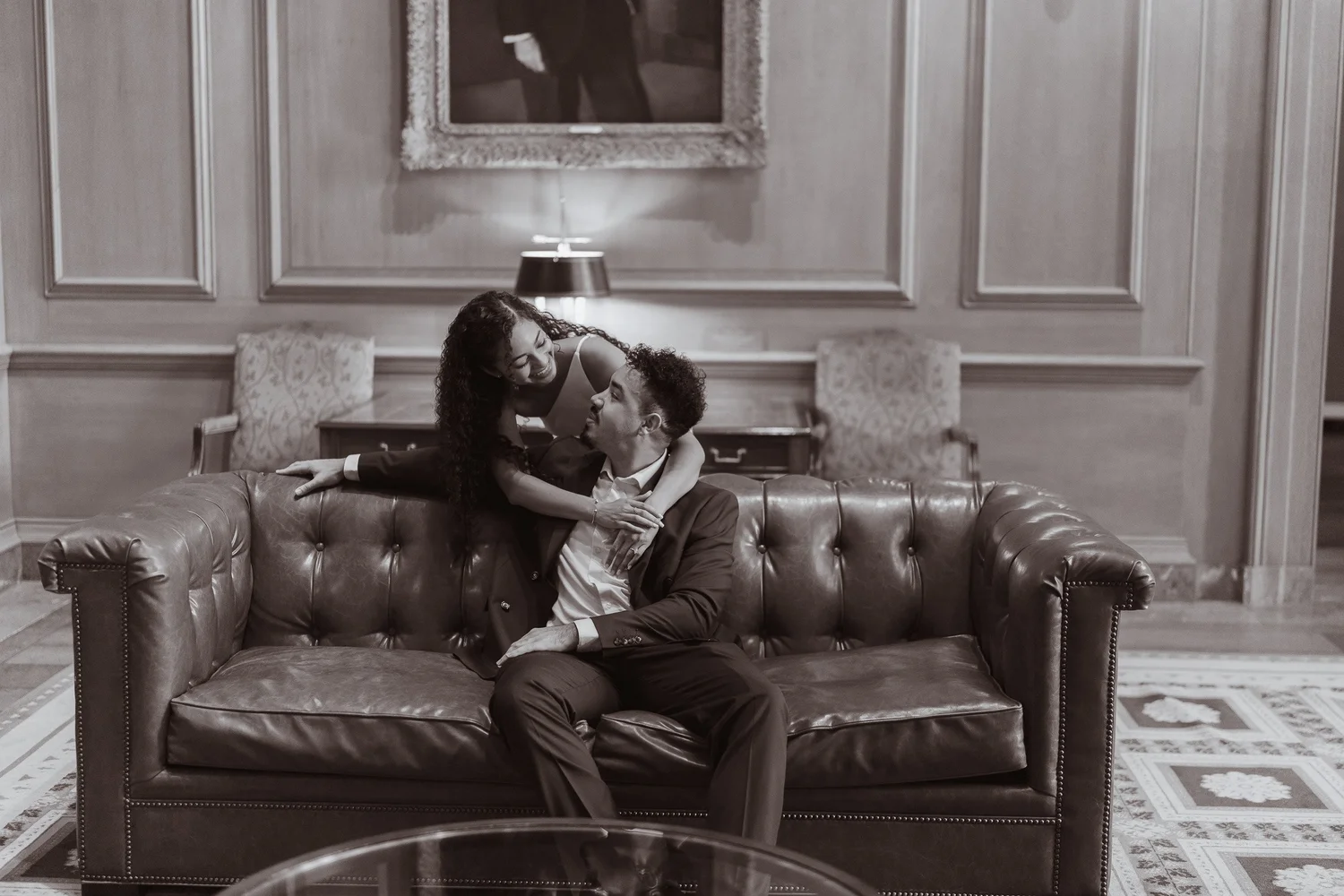 Black-and-white photograph of a couple sharing a playful moment on a leather sofa.