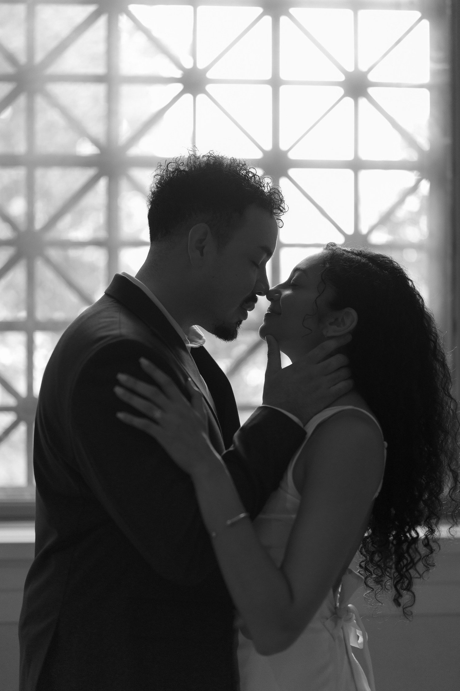 Black-and-white close-up of a couple leaning together by a patterned window.