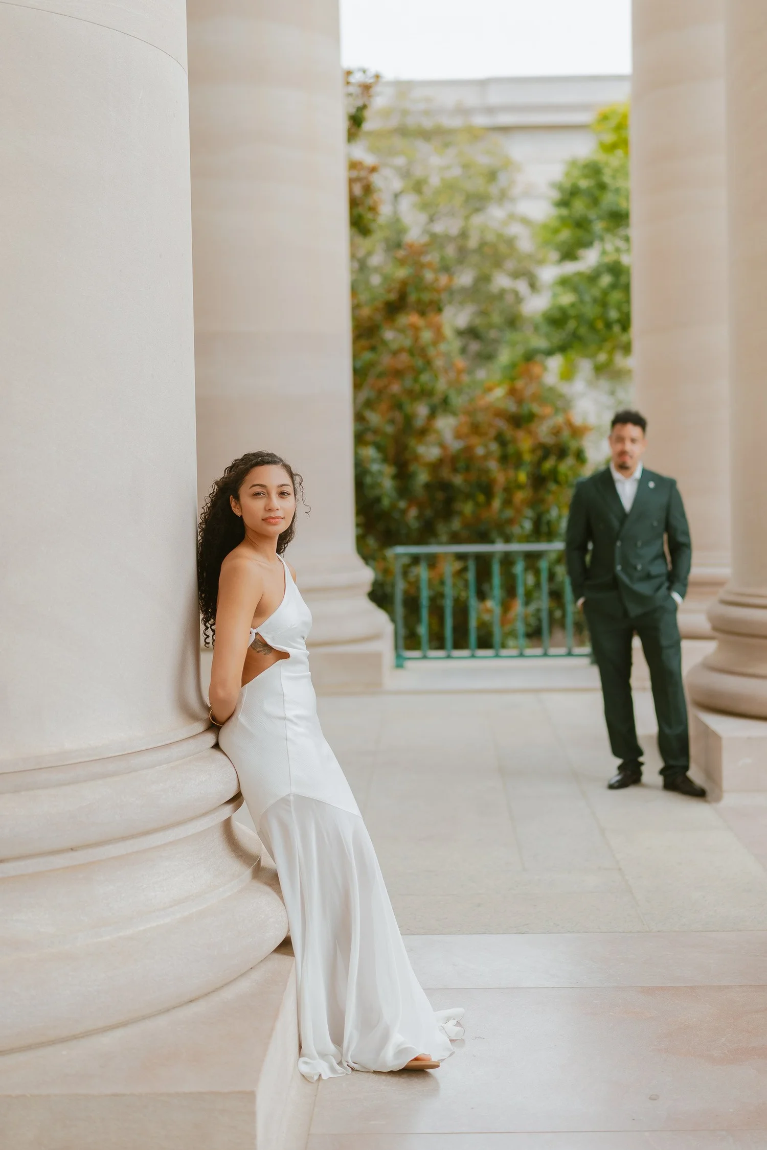 A woman beside a pale column with her partner softly in the background.