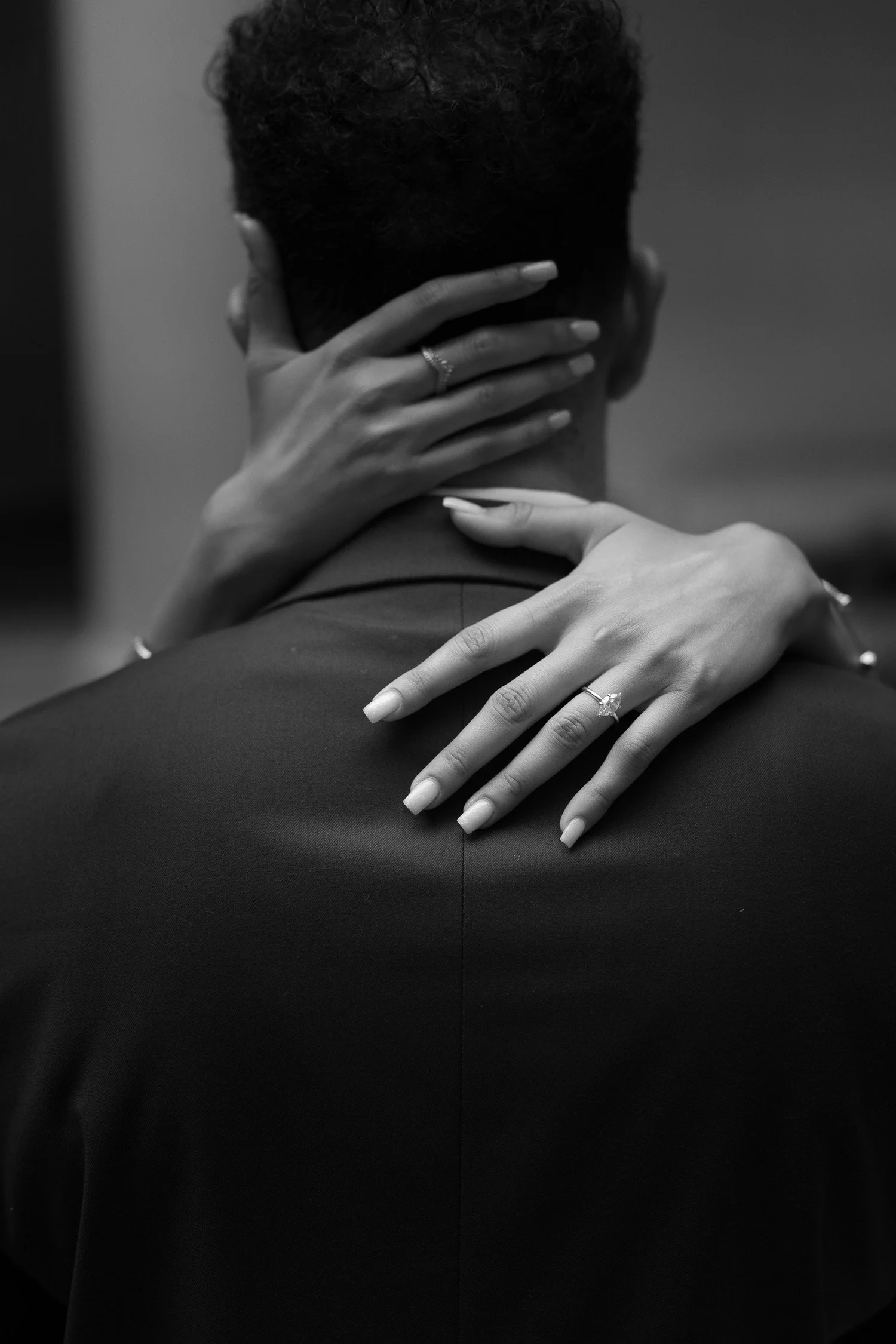 Black-and-white detail of an engagement ring and hands around a partner's shoulders.