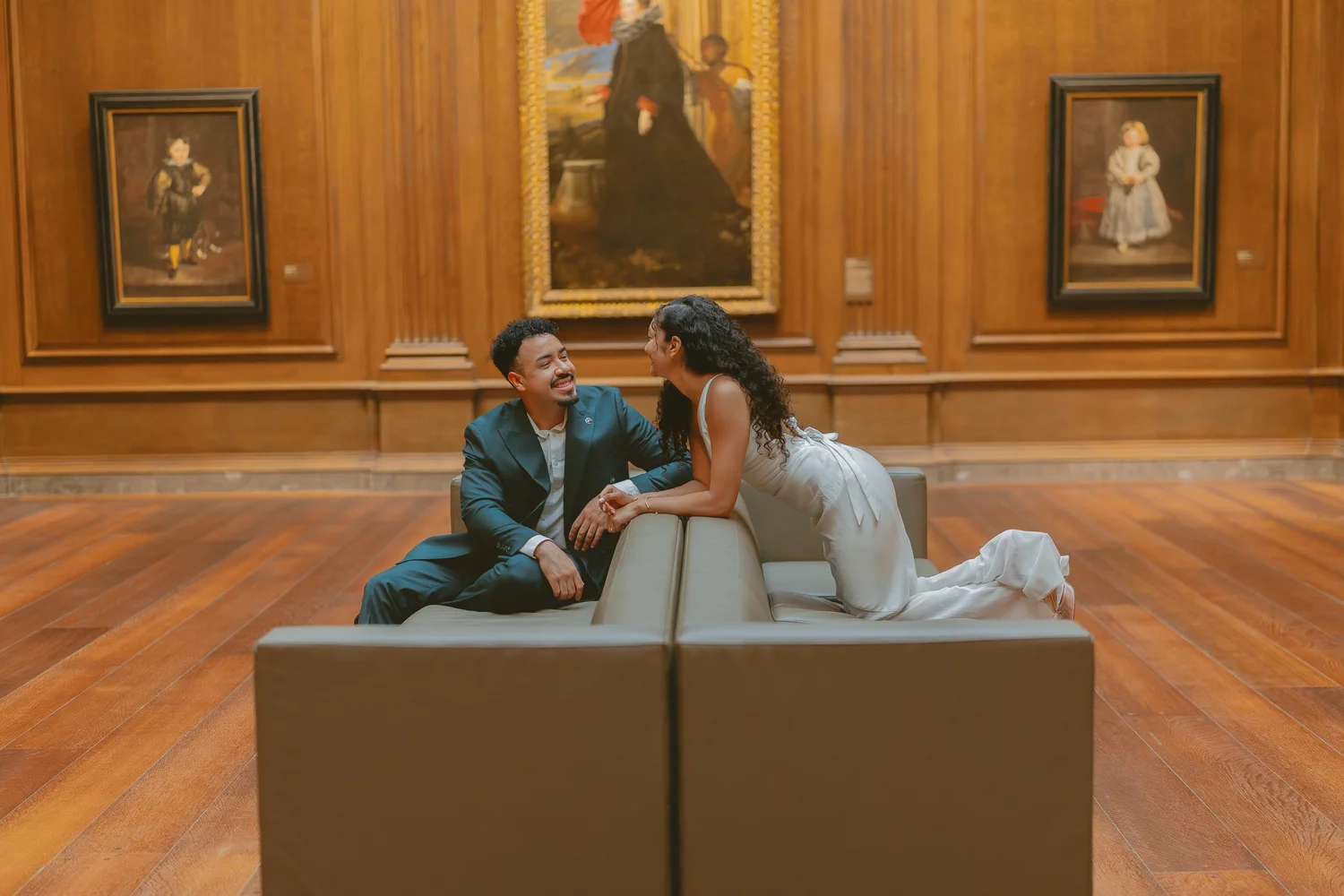 Couple laughing together on a bench in a warm wood-paneled art gallery.