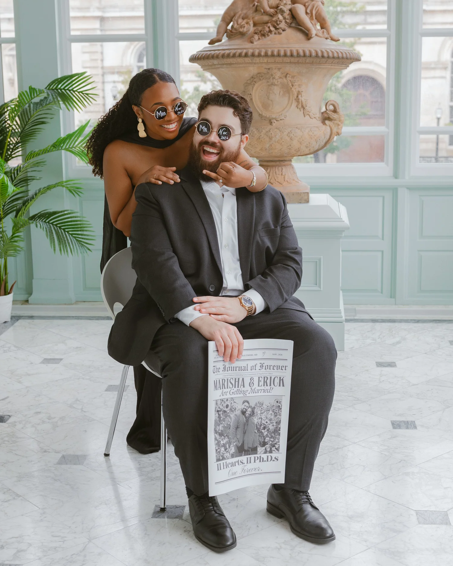 Couple laughing in playful sunglasses beside a sculpture and bright windows.