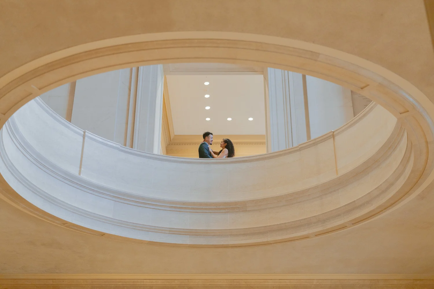 Couple framed by the oval opening of a sweeping marble staircase.