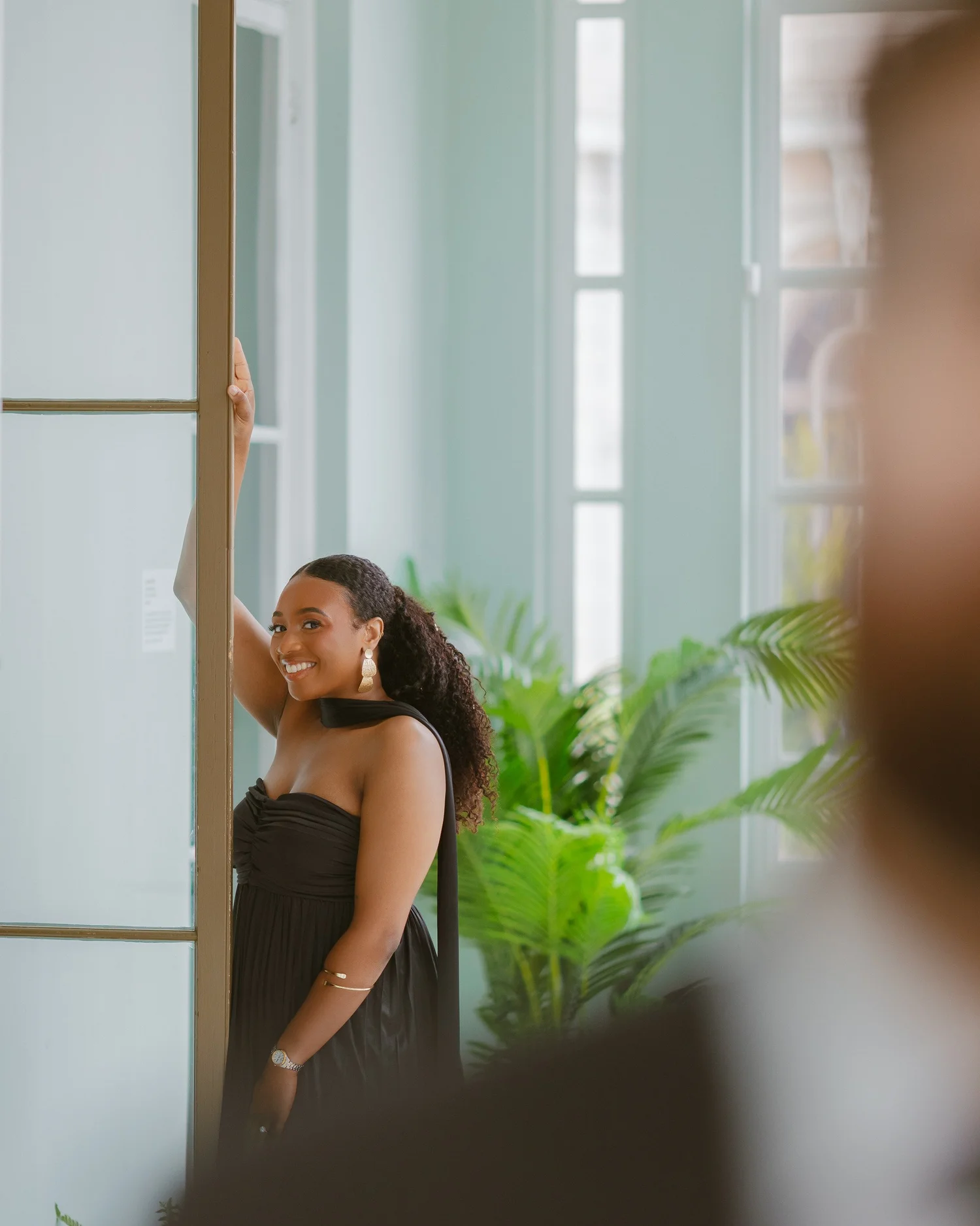 A woman smiling beside a tall window and green plants.