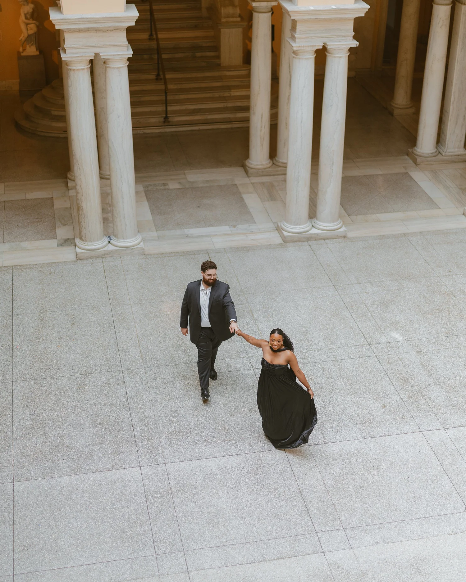 Couple walking hand in hand across a stone courtyard beneath arches.