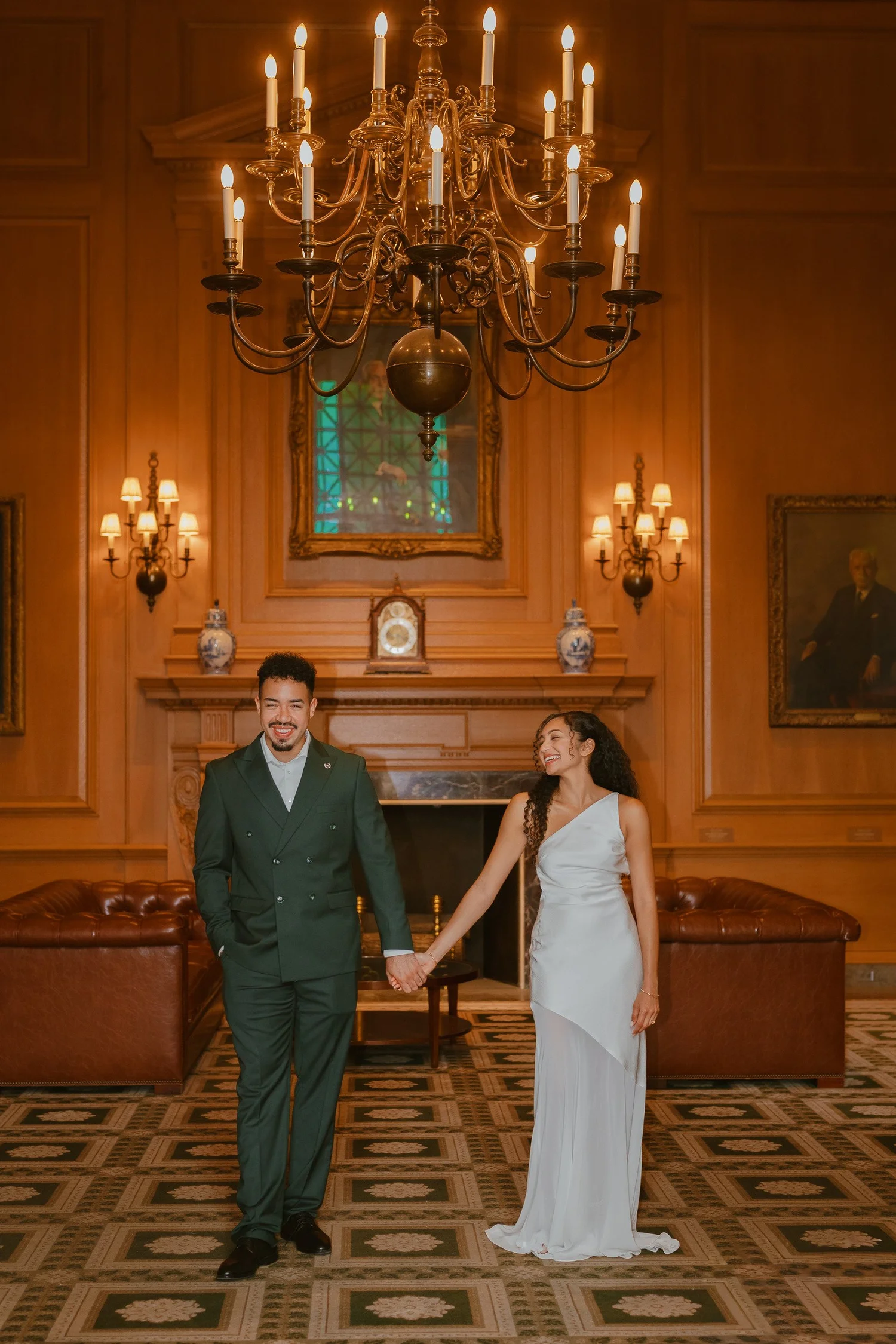 Couple holding hands in a wood-paneled room beneath a chandelier.