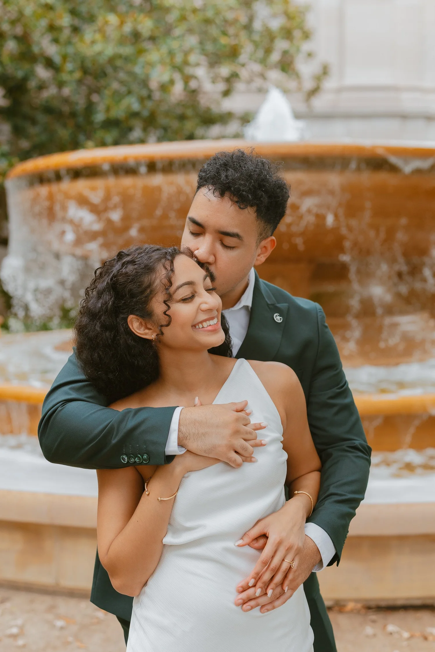 A man kissing his smiling partner's temple beside a tiered fountain.