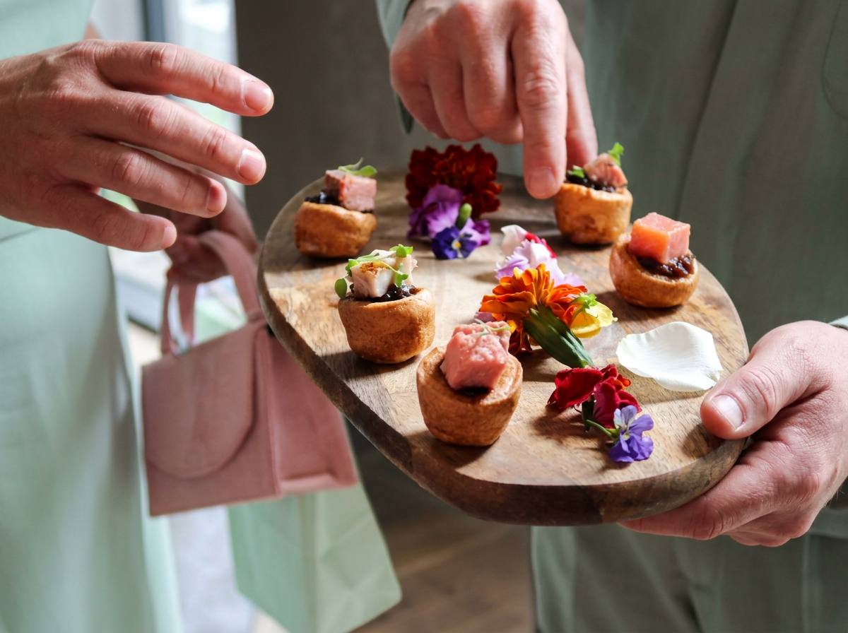 A wooden board of small Yorkshire pudding canapés topped with rare beef and edible flowers, held by two hands at a Taste Cuisine event