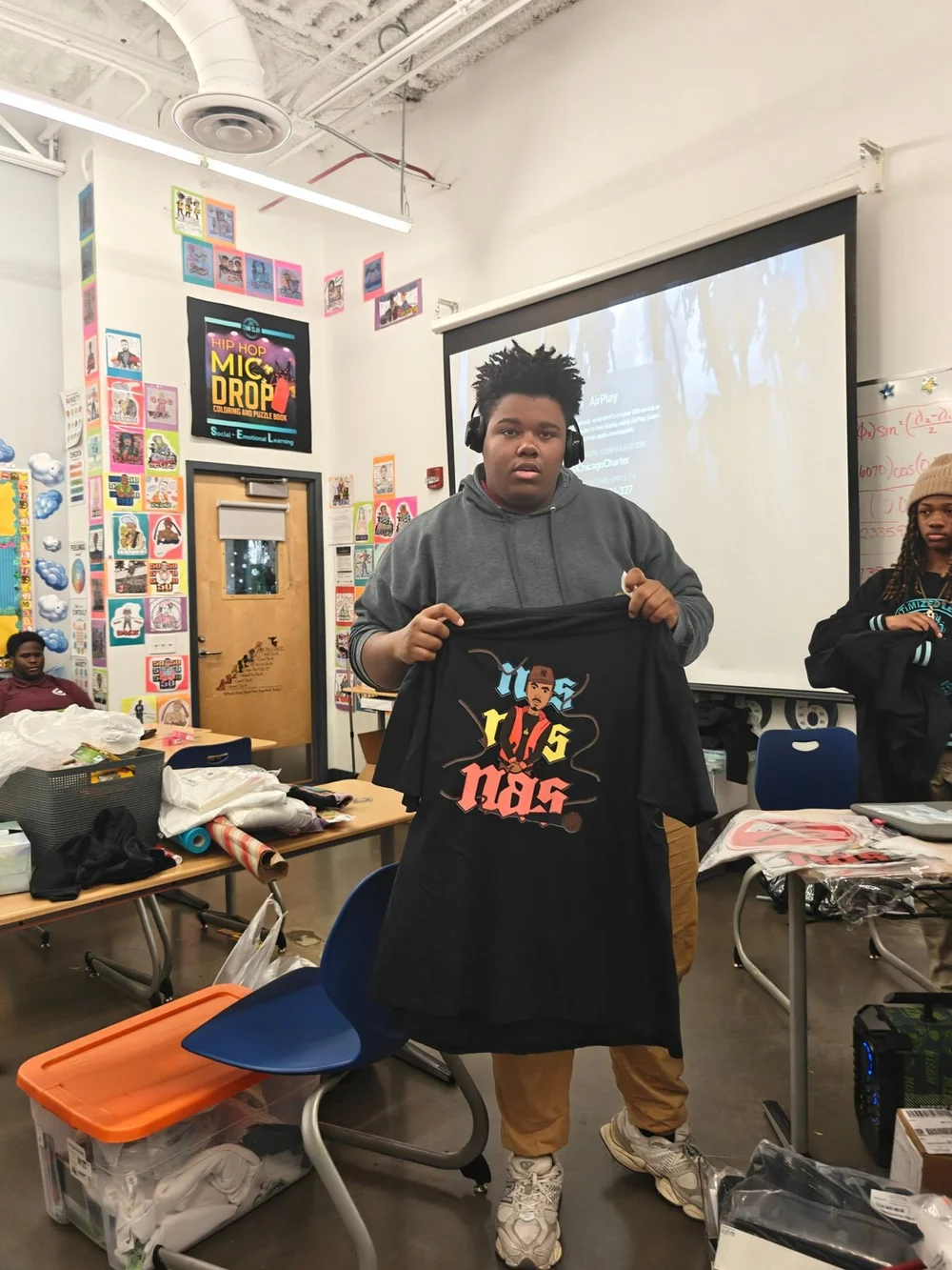 A student holds up a t-shirt he designed, with a Hip Hop Mic Drop poster behind him