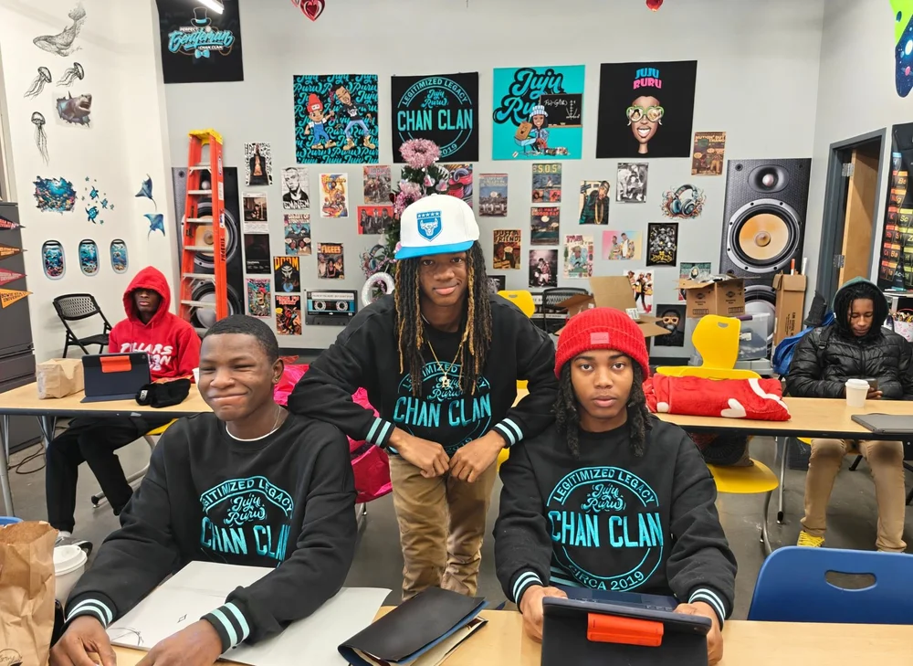 Three students in Chan Clan sweatshirts at their desks with notebooks and tablets