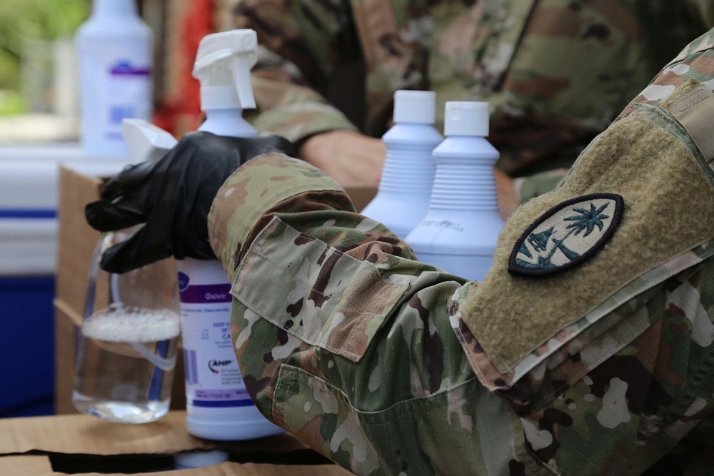 Gloved hands holding a spray bottle of disinfectant next to cleaning supplies